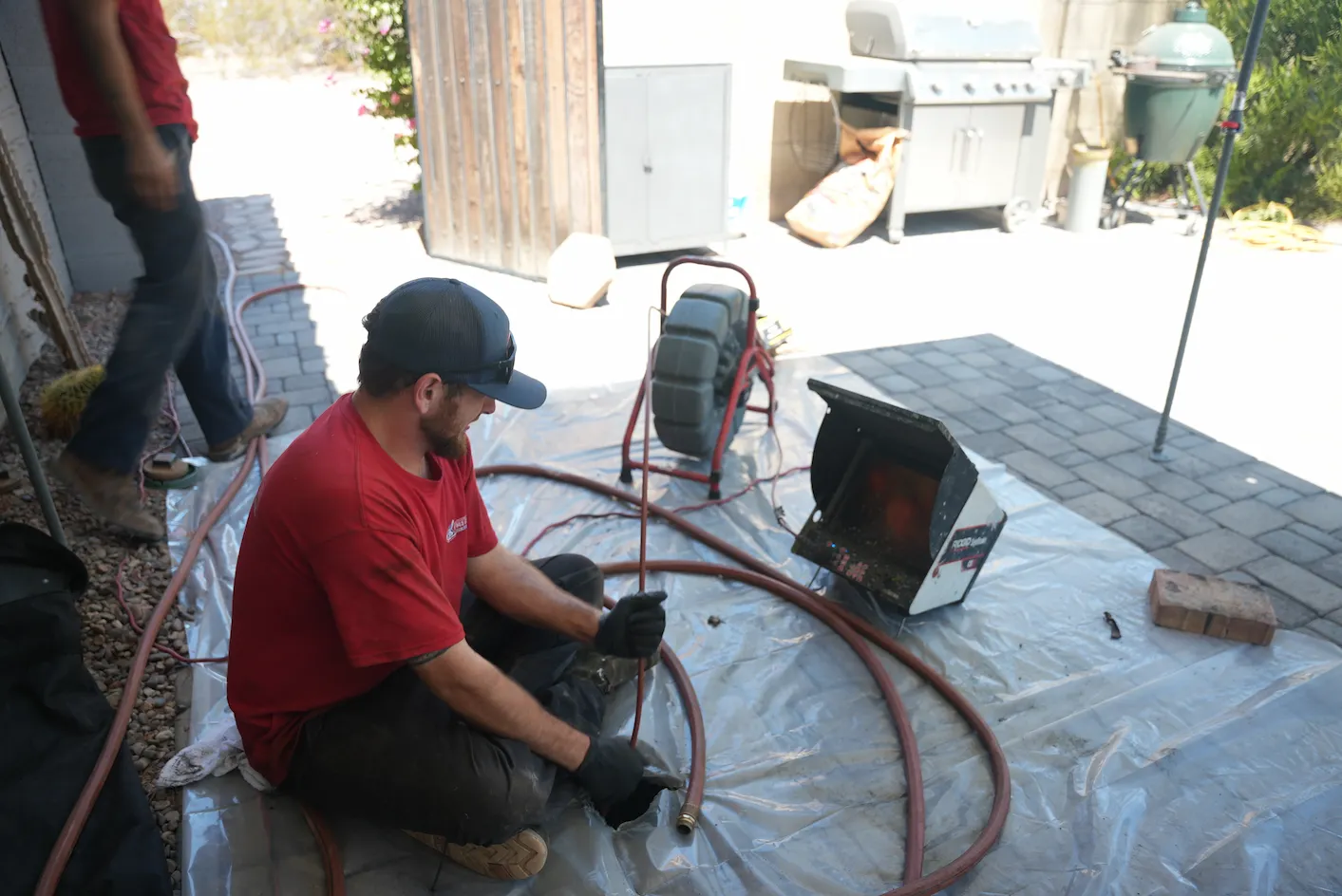 A male plumber wearing a red shirt, dark pants, and a baseball cap is seated on a clear plastic tarp, feeding a flexible cable into a drain inspection camera system.