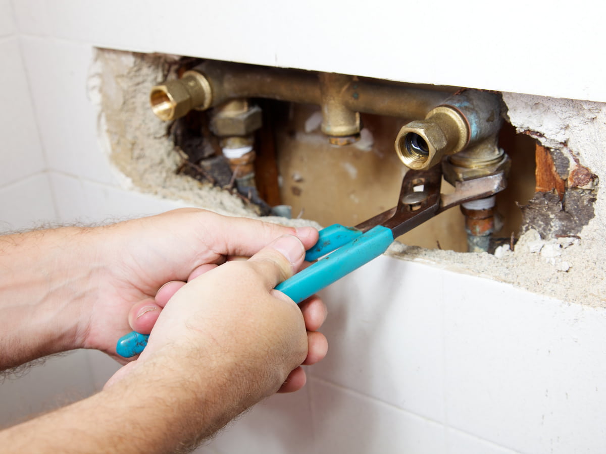 Plumber repairing an indoor water and sewer line behind a wall using a wrench.